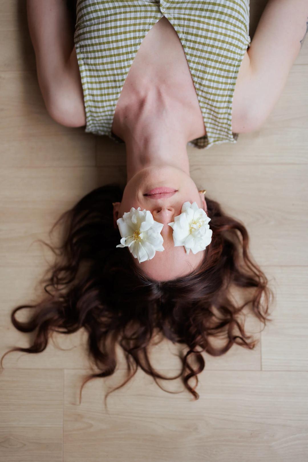 Une femme allongée sur le parquet, des fleurs blanches posées sur les yeux